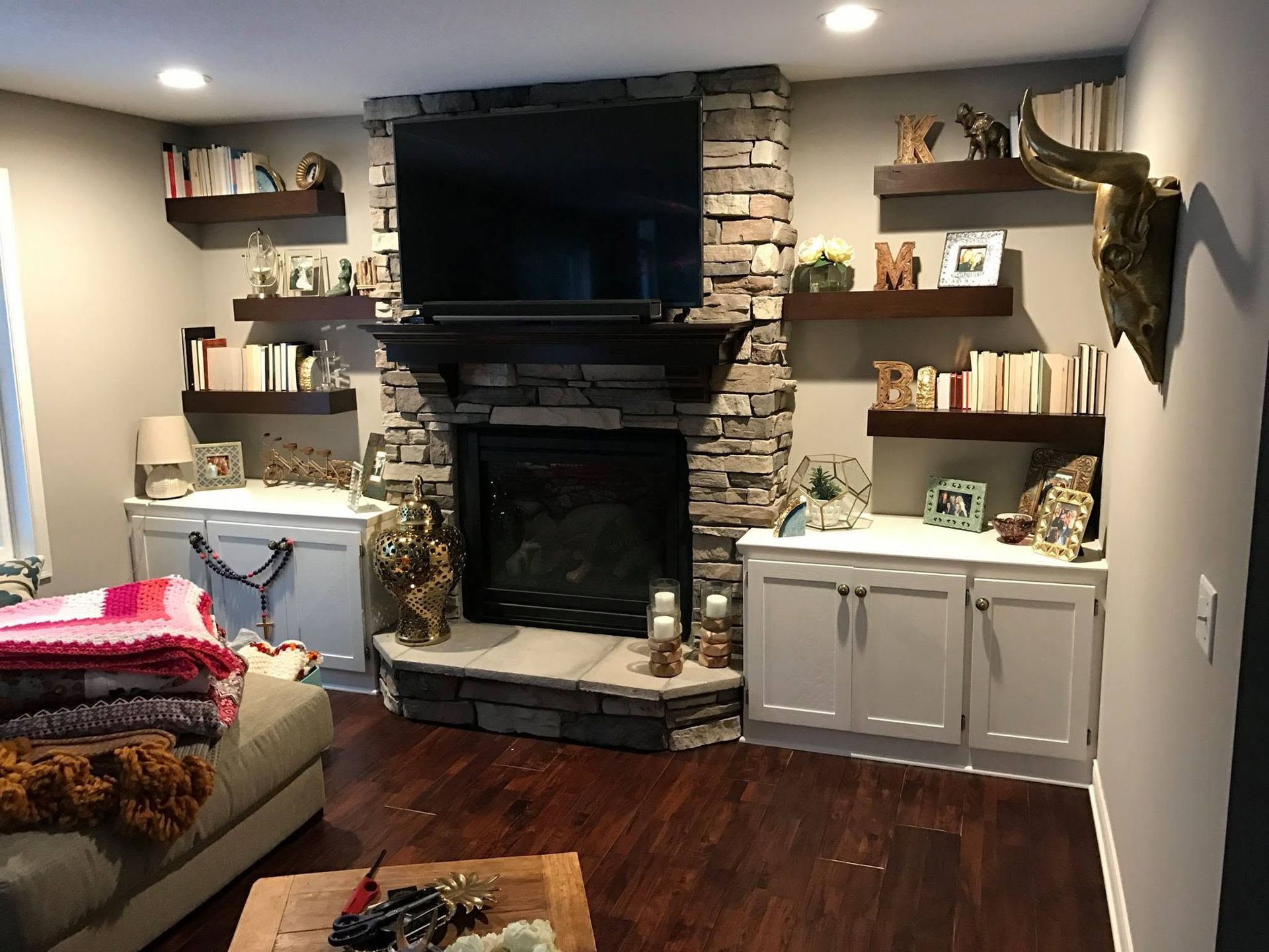Cozy living room with a fireplace, TV, wall shelves, and wood floors, lit by recessed ceiling lights
