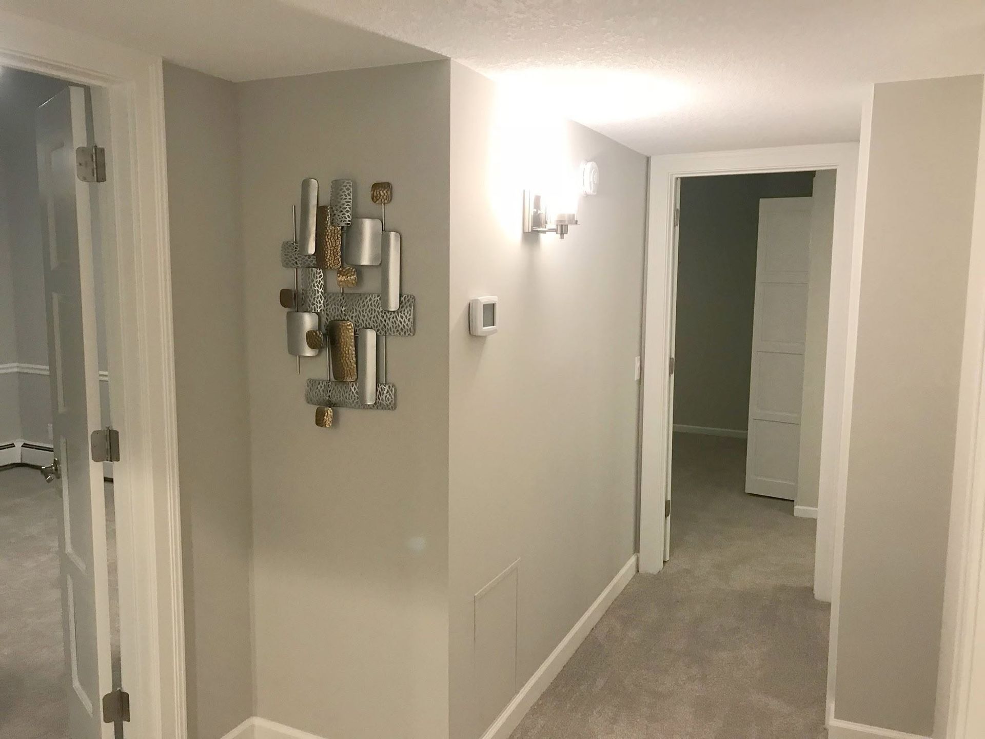 Empty beige hallway with carpet, wall decor, and a lit doorway at the far end