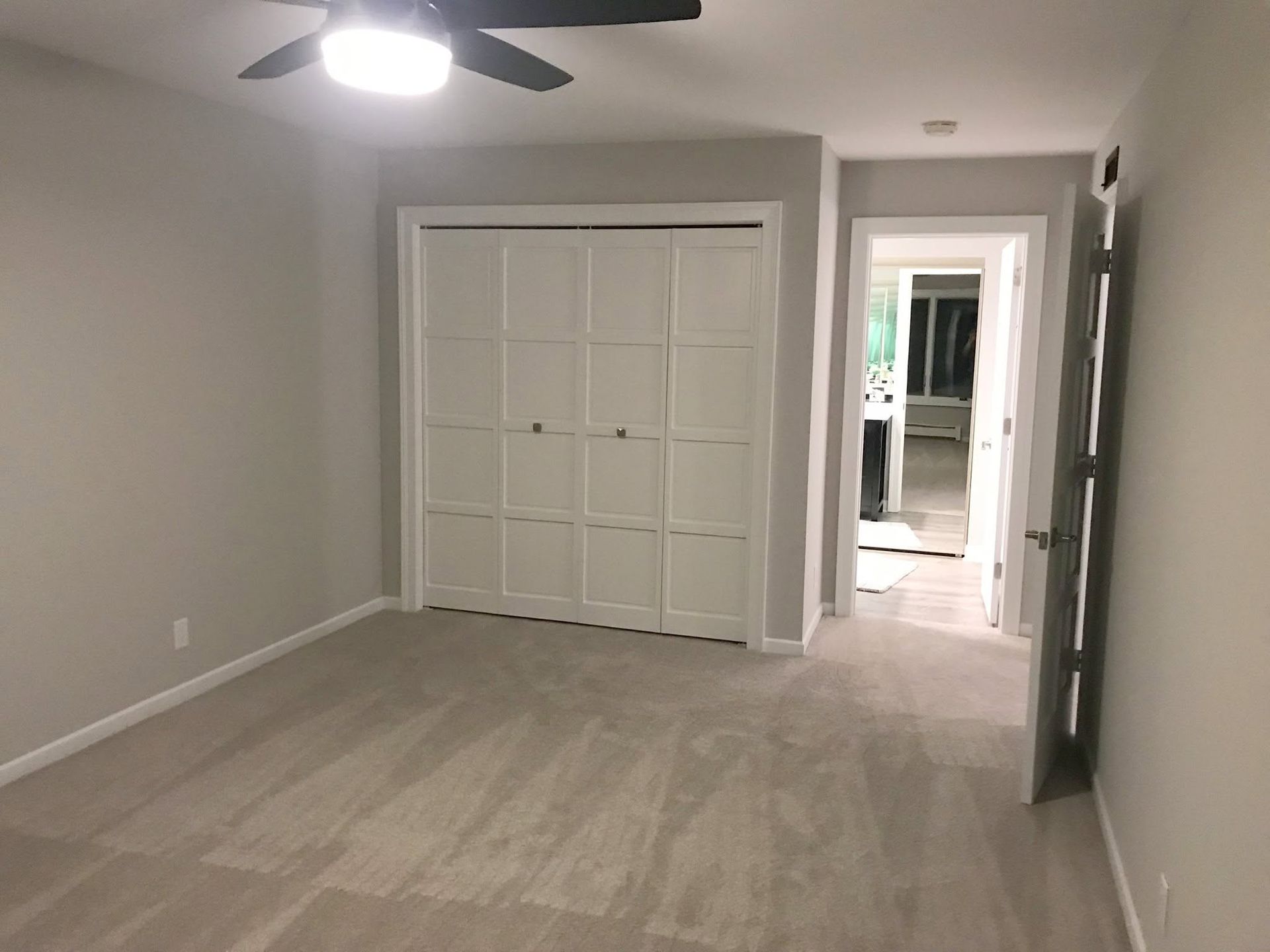 Empty gray bedroom with ceiling fan, white closet doors, and doorway to a bright hall