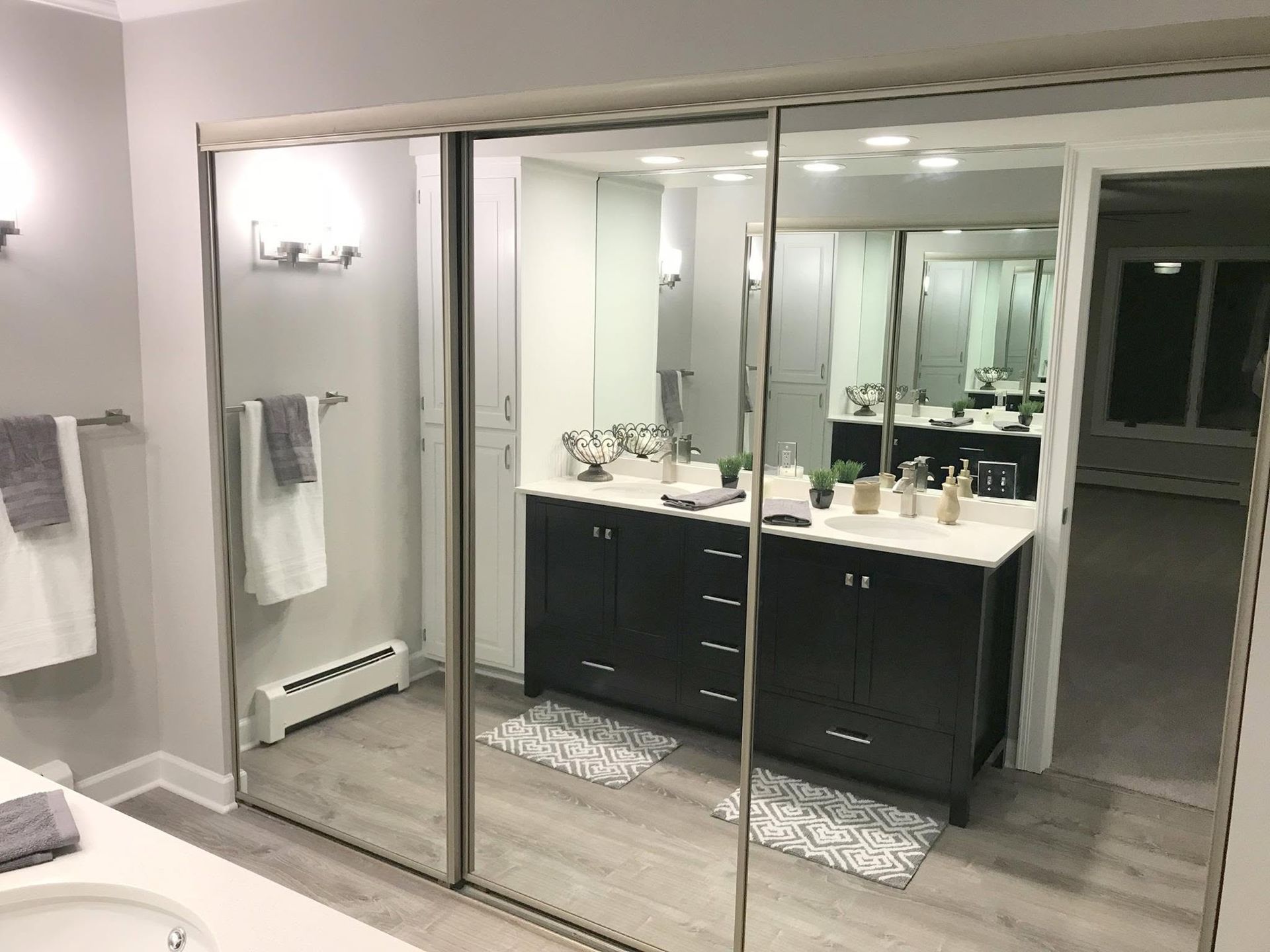 Modern bathroom with double vanity, large mirrored closet doors, and light wood flooring