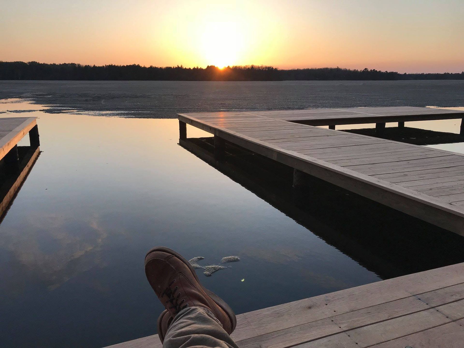 Boot on dock beside calm lake at sunset, with orange sky reflected on the water.
