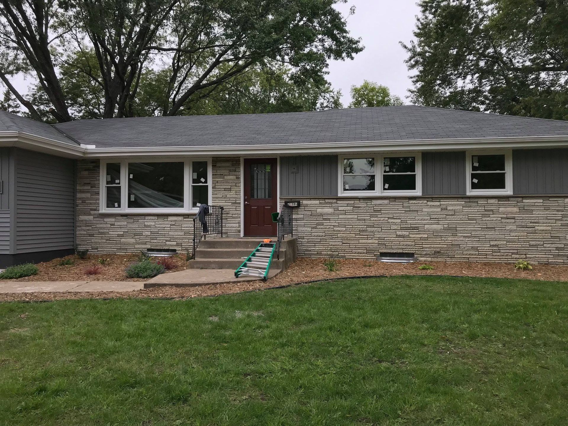 Single-story house with stone facade, gray siding, red door, and front lawn under trees