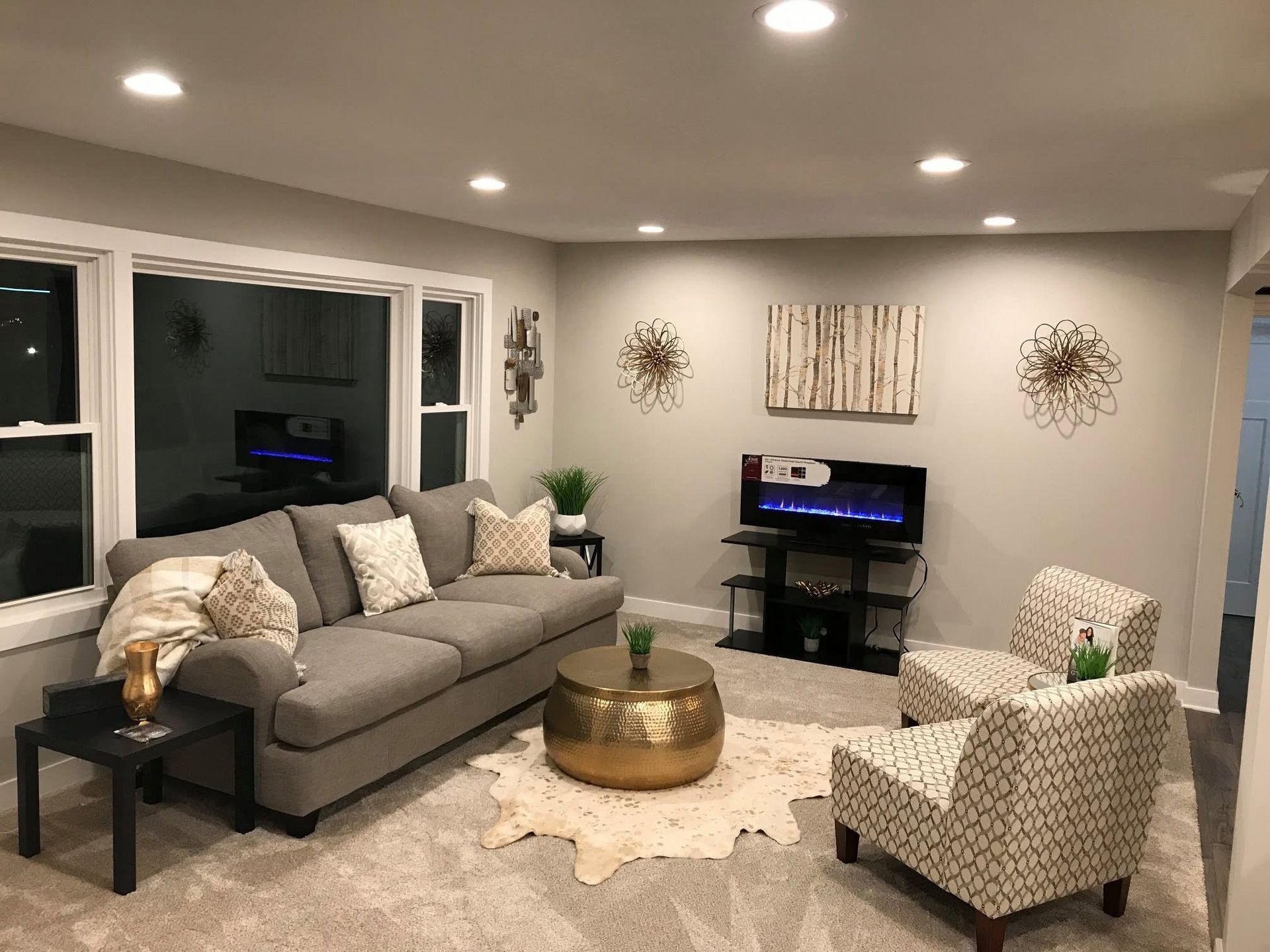 Bright living room with gray sectional sofa, patterned chairs, TV, round coffee table, and recessed lighting.