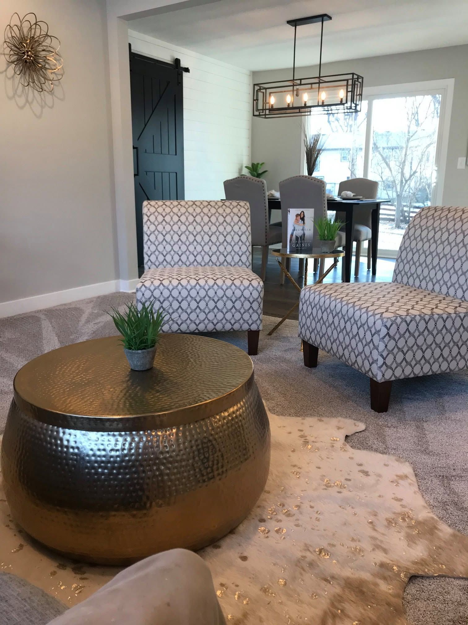 Modern living room with patterned chairs, bronze coffee table, dining area, and large windows with dark curtains.