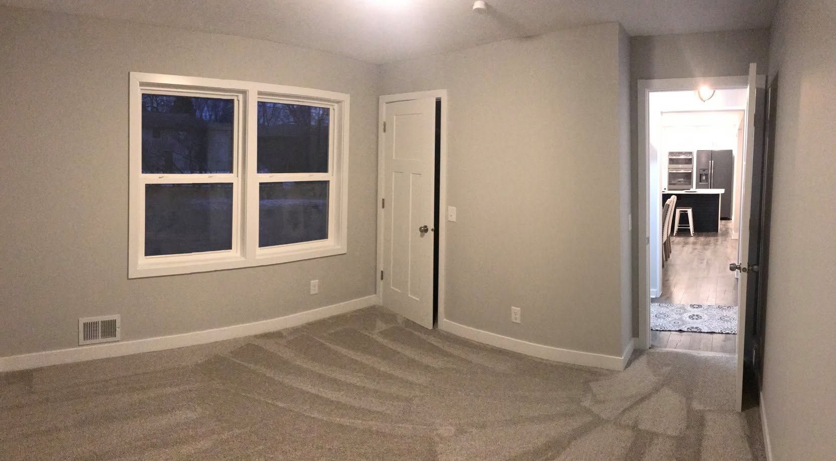 Empty beige room with carpet, two windows, and an open doorway to a lit kitchen.