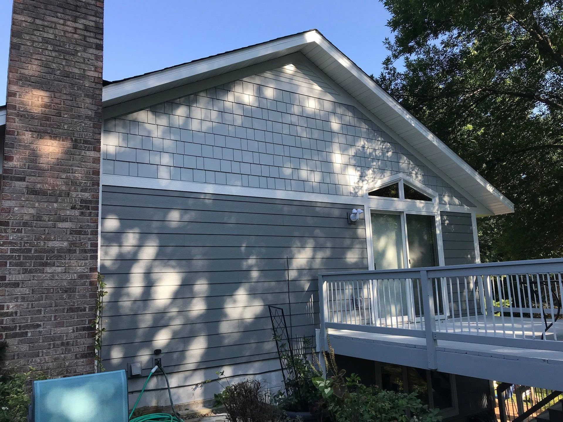Exterior of a house with gray siding, brick chimney, and a white-railed deck in dappled sunlight