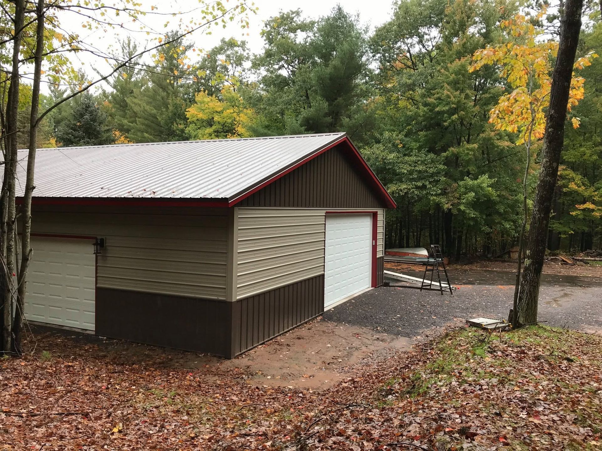 Small beige and brown shed with a white roof in a wooded yard covered in fallen leaves
