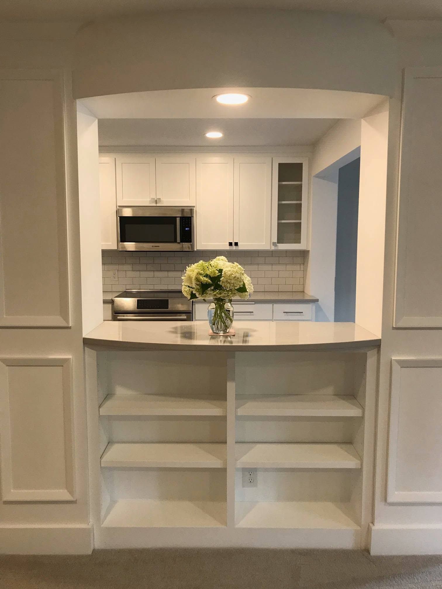 White kitchen pass-through with shelves, counters, stainless appliances, and a vase of flowers.