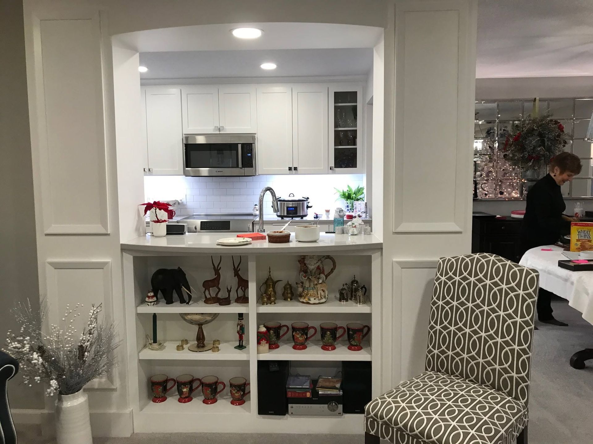 White kitchen pass-through with shelves of figurines and a patterned chair; person standing in adjacent room