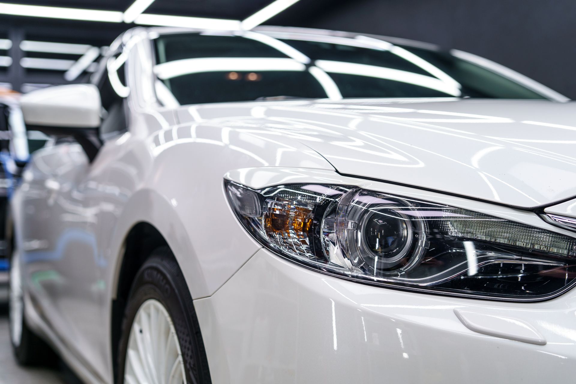 Close-up of a white car’s front end and headlight under bright detailing shop lighting.