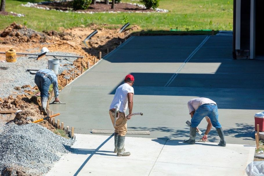Three workers smoothing fresh concrete on a driveway slab beside a house construction site