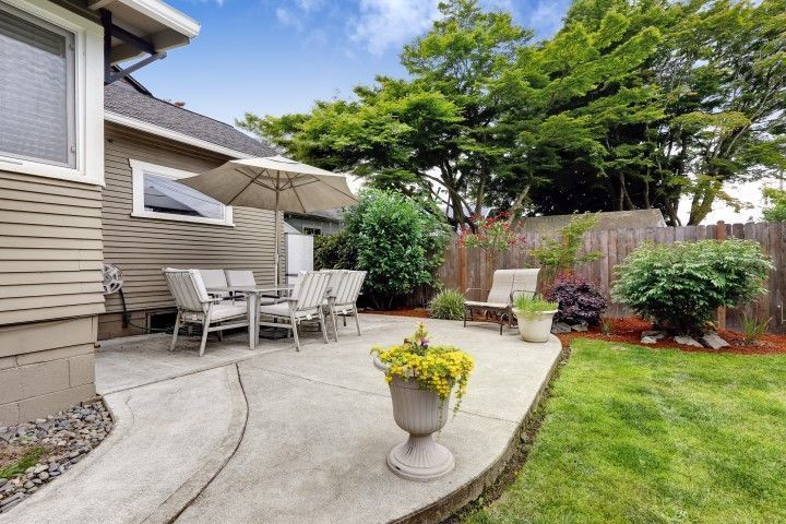 Patio with outdoor table and chairs beside a house, potted flowers, and a green lawn on a sunny day