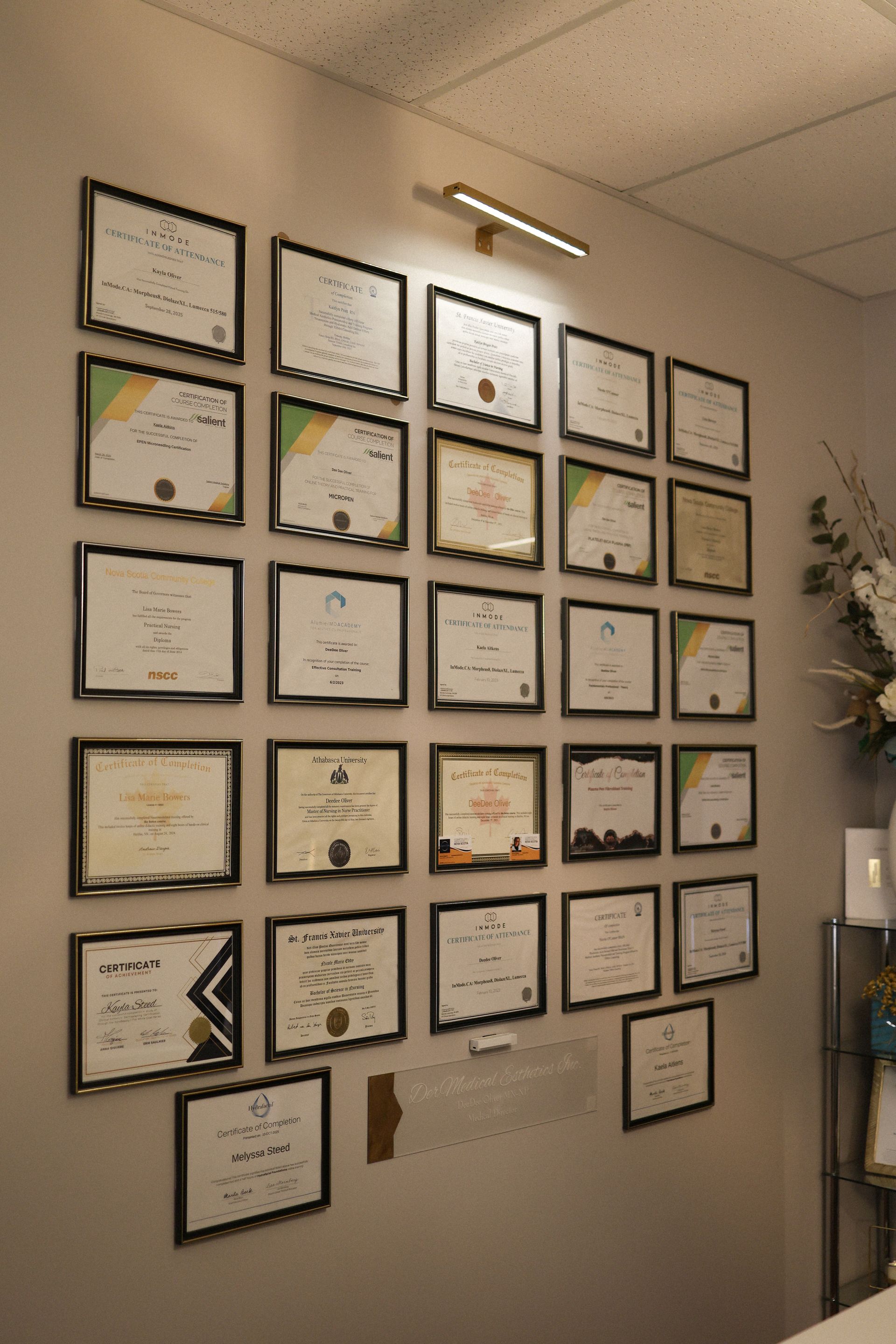 Wall displaying numerous framed certificates in a professional setting, lit by a small overhead light.
