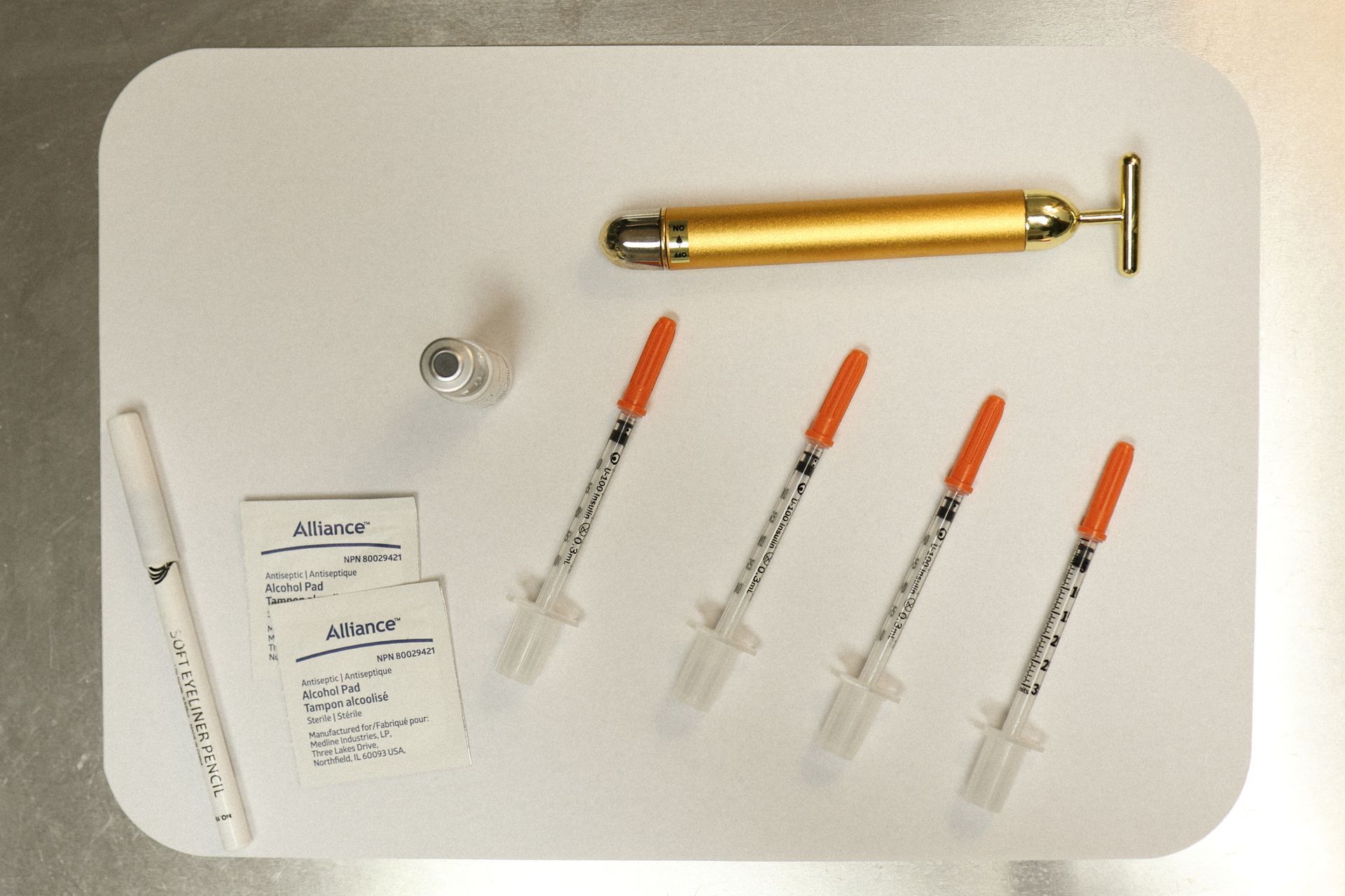 White tray with syringes, a pen, small cards, a vial, and a golden face massager.