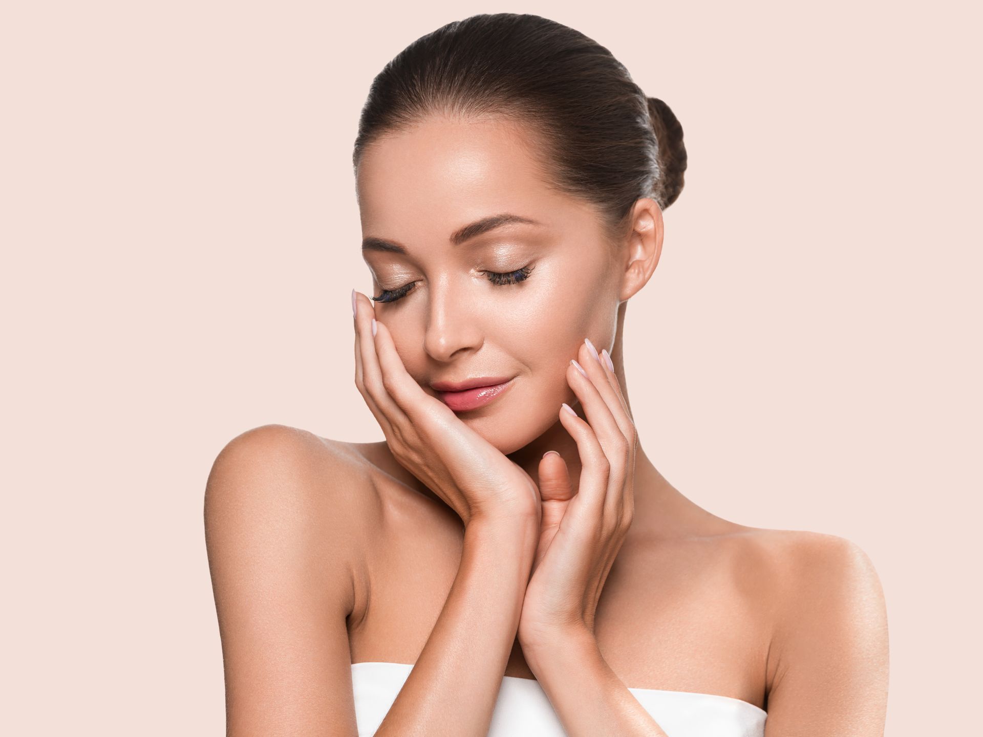 Woman with glowing skin, eyes closed, touching her face with both hands against a light tan background.
