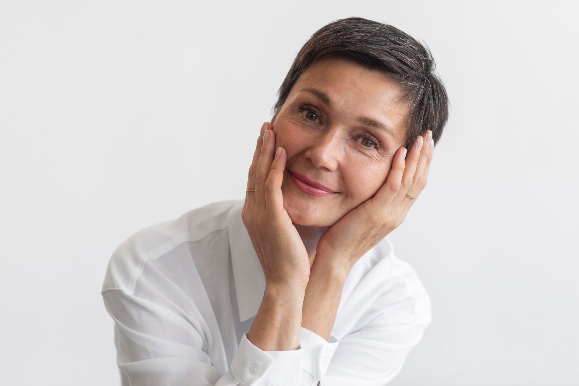 Woman with short dark hair, smiles gently, hands on her face. She wears a white shirt, against a white background.