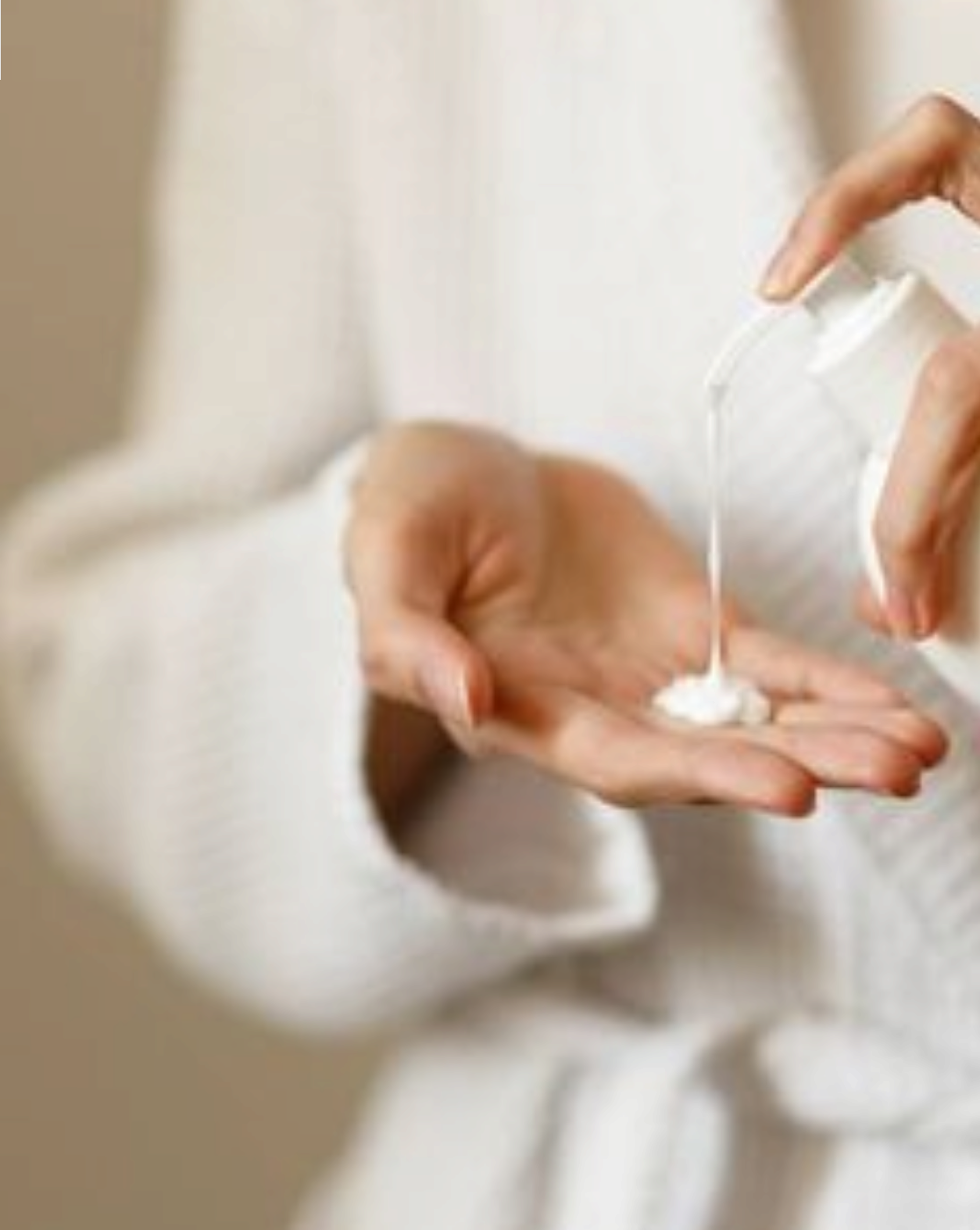 Woman dispensing white lotion from a bottle into her palm while wearing a white robe.
