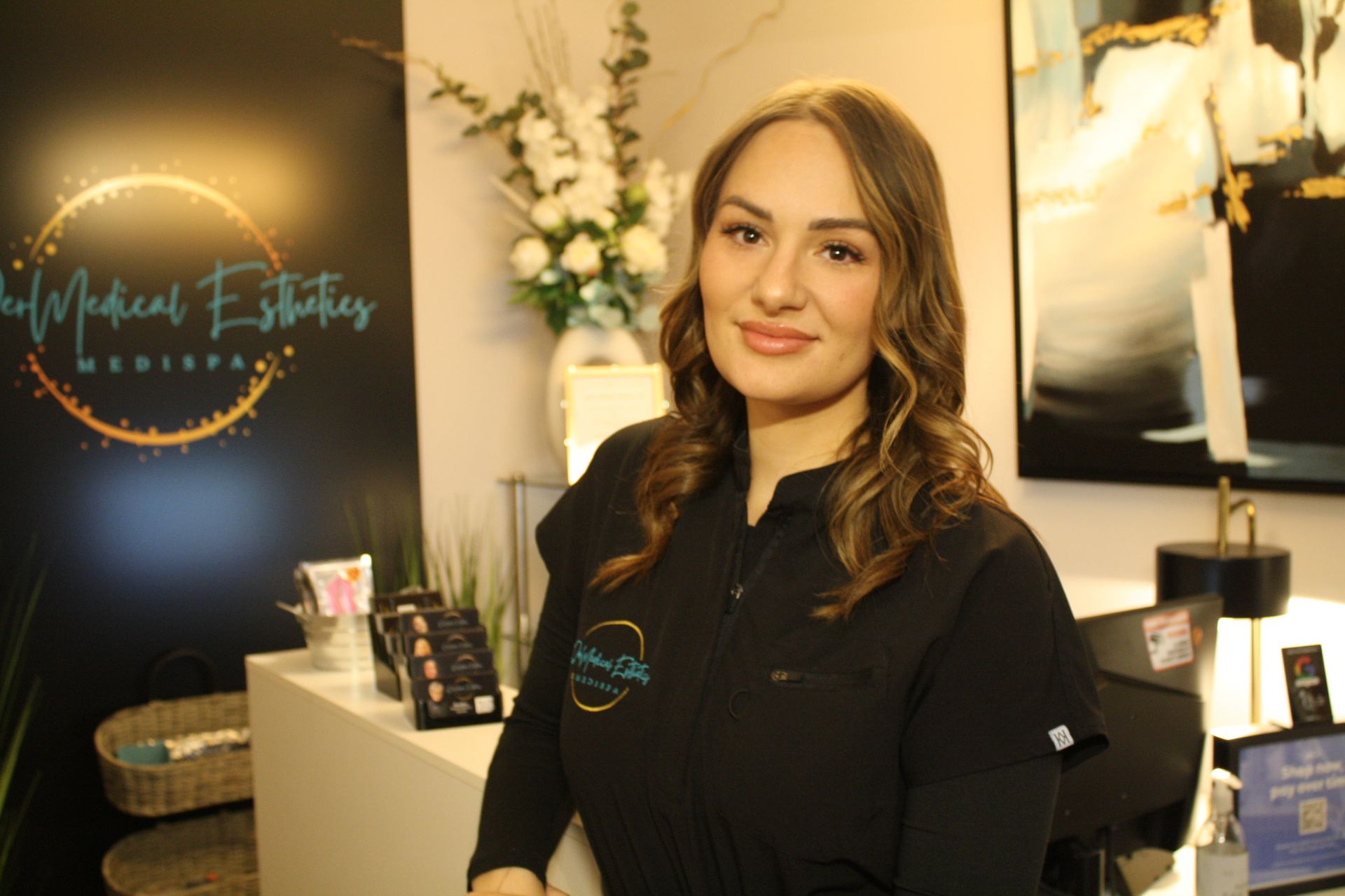 Woman in black scrubs smiling near a medical spa reception desk, logo in background.