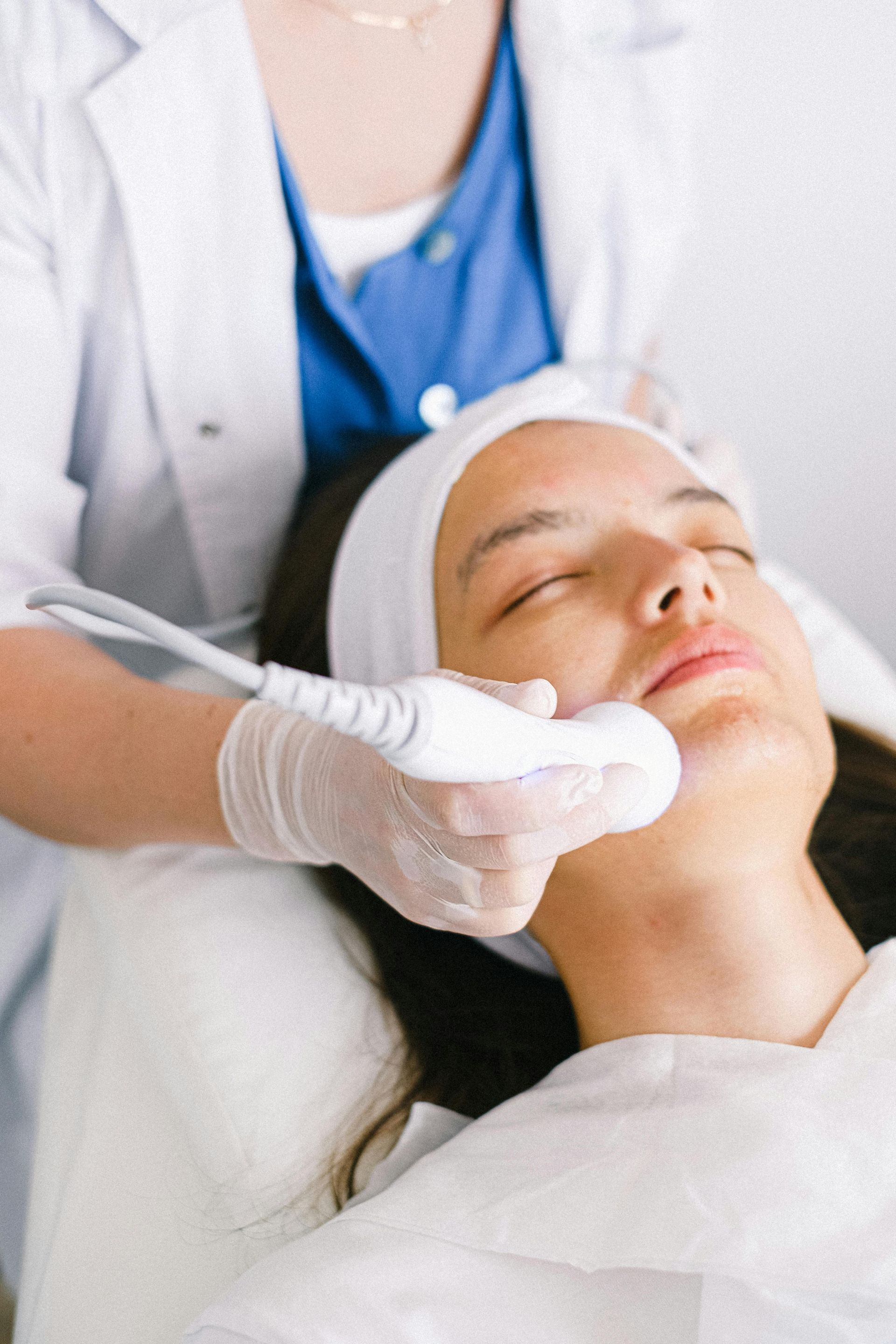 Woman receiving facial treatment, lying down with eyes closed; hands massaging face with cream.