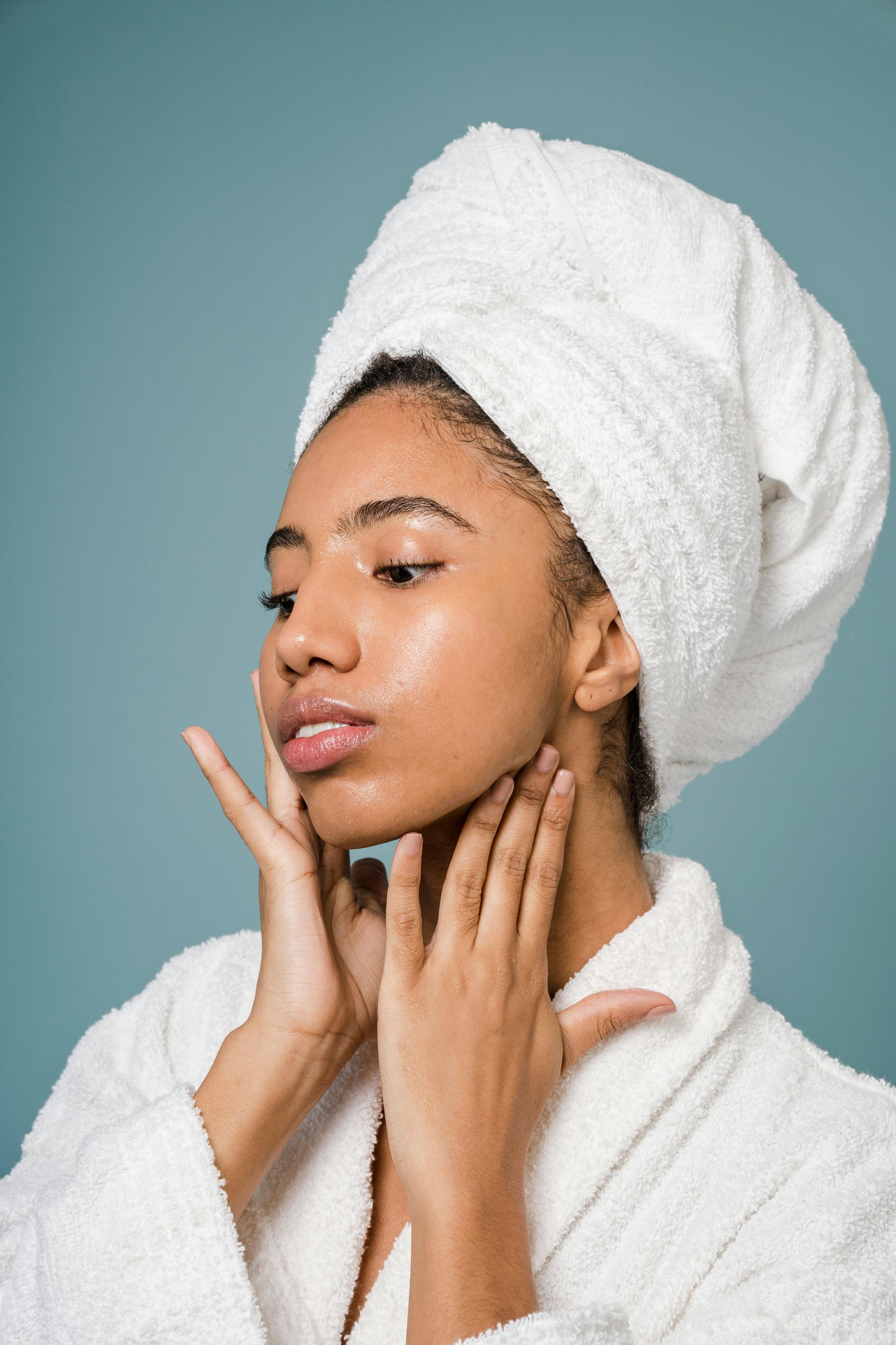 Woman with wet, glowing skin wrapped in a white towel and robe, touching her neck with hands.