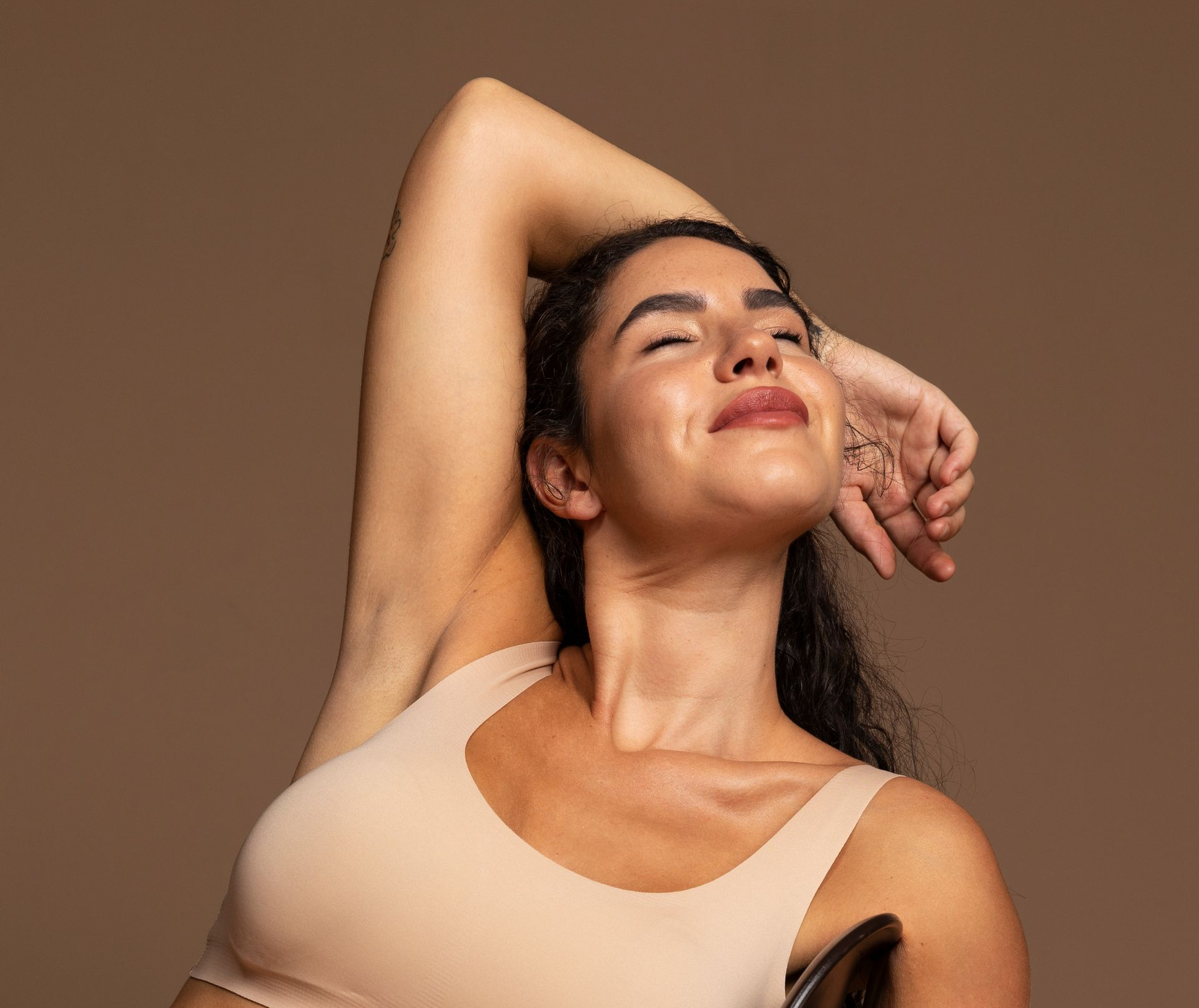 Woman with dark hair, arms raised, smiling against a brown backdrop. She wears a beige top.