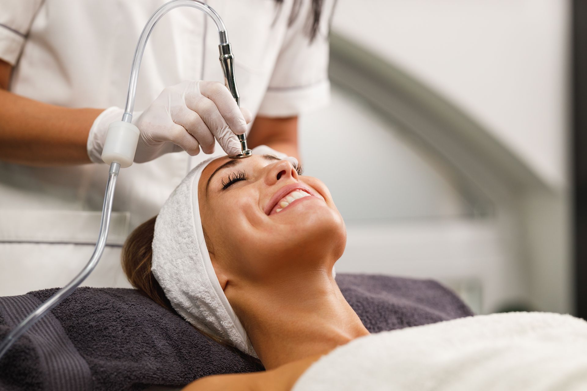Woman receiving facial treatment, smiling. Aesthetican uses a skin resurfacing device in a bright, indoor setting.