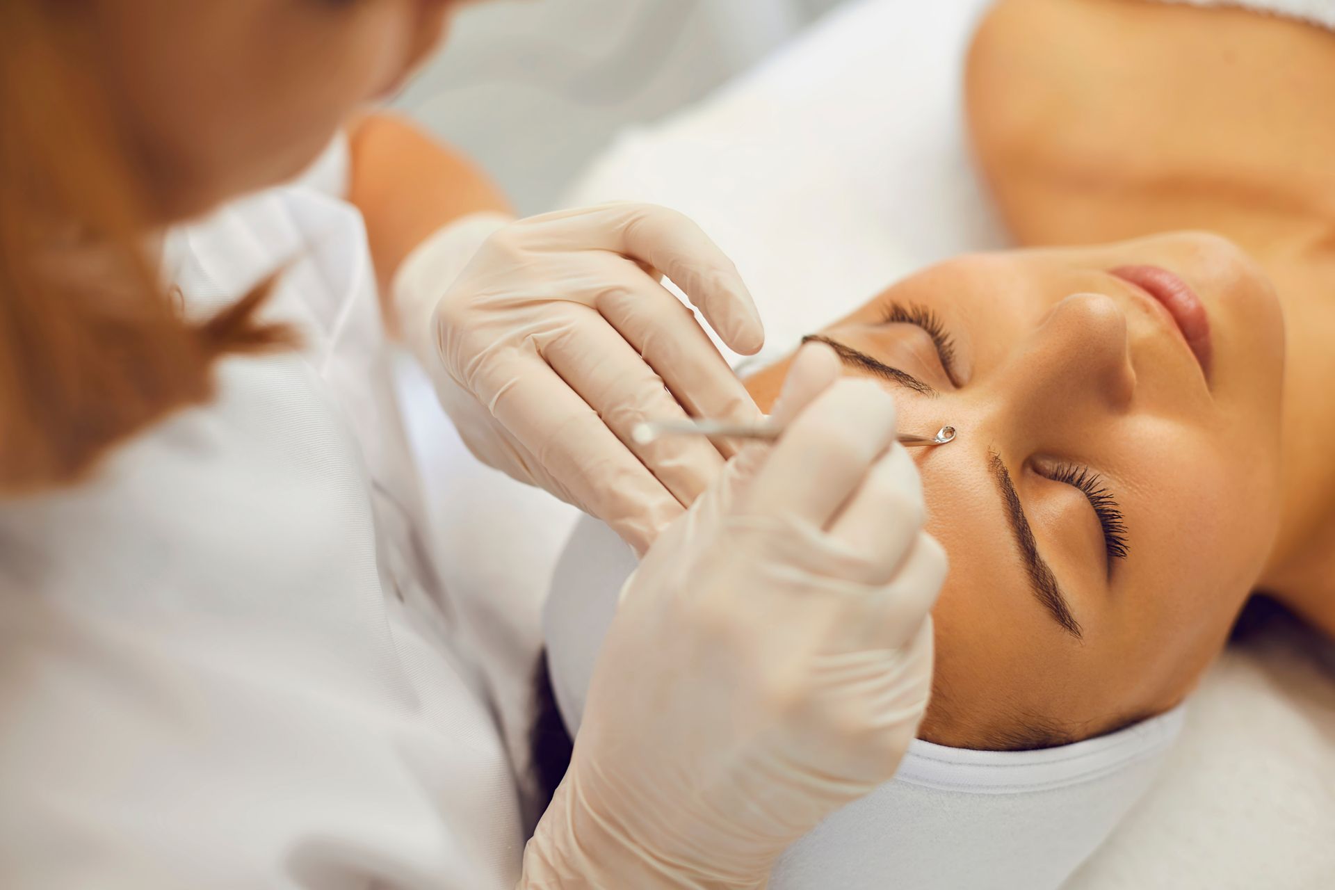 Aesthetician wearing gloves removing eyebrow hair with tweezers from a reclining client