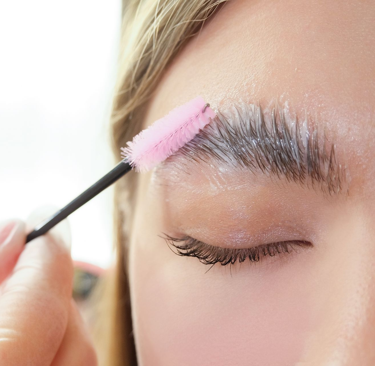 A person brushing their eyebrow with a pink spoolie applicator, applying a brow styling product.