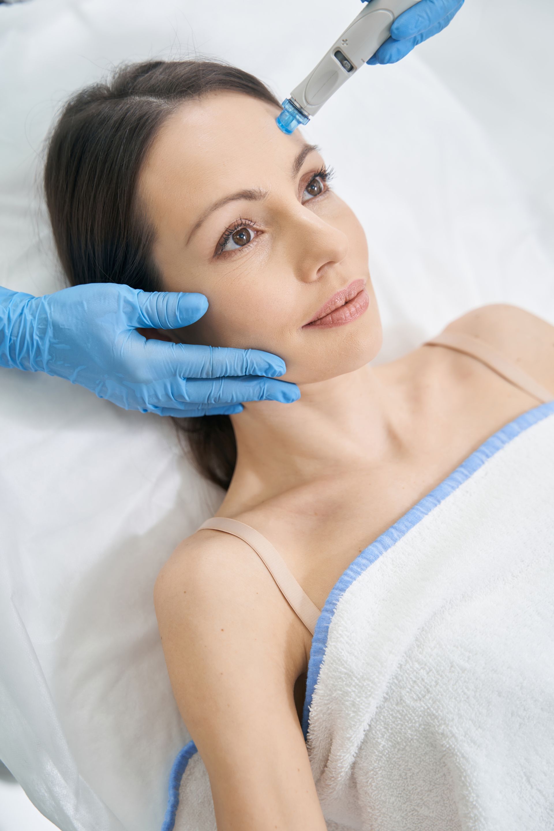 Woman receiving facial treatment; blue-gloved hands holding device and supporting face, white towel, spa setting.