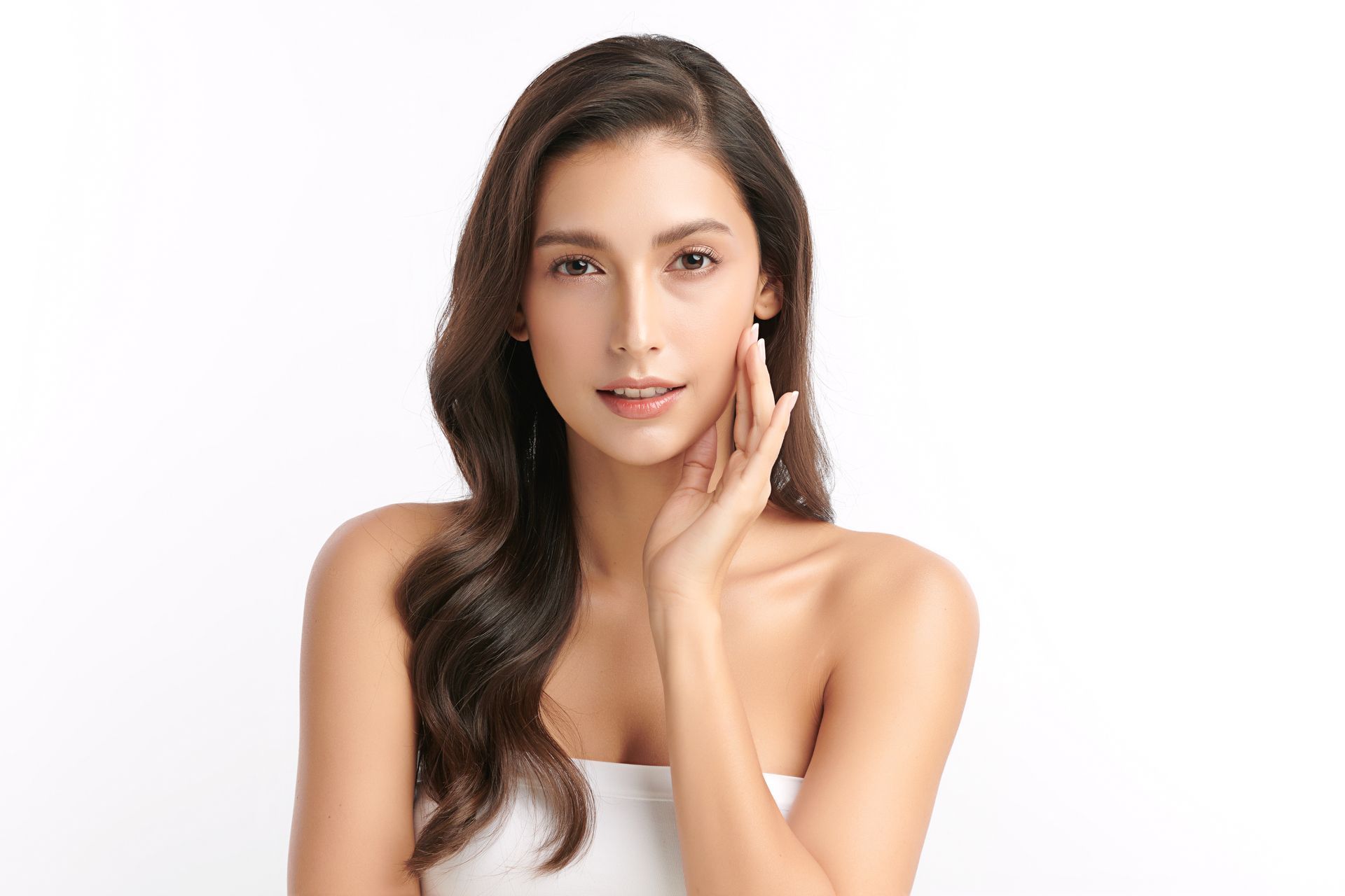Woman with wavy brown hair, looking at the viewer, touching her face, white background.