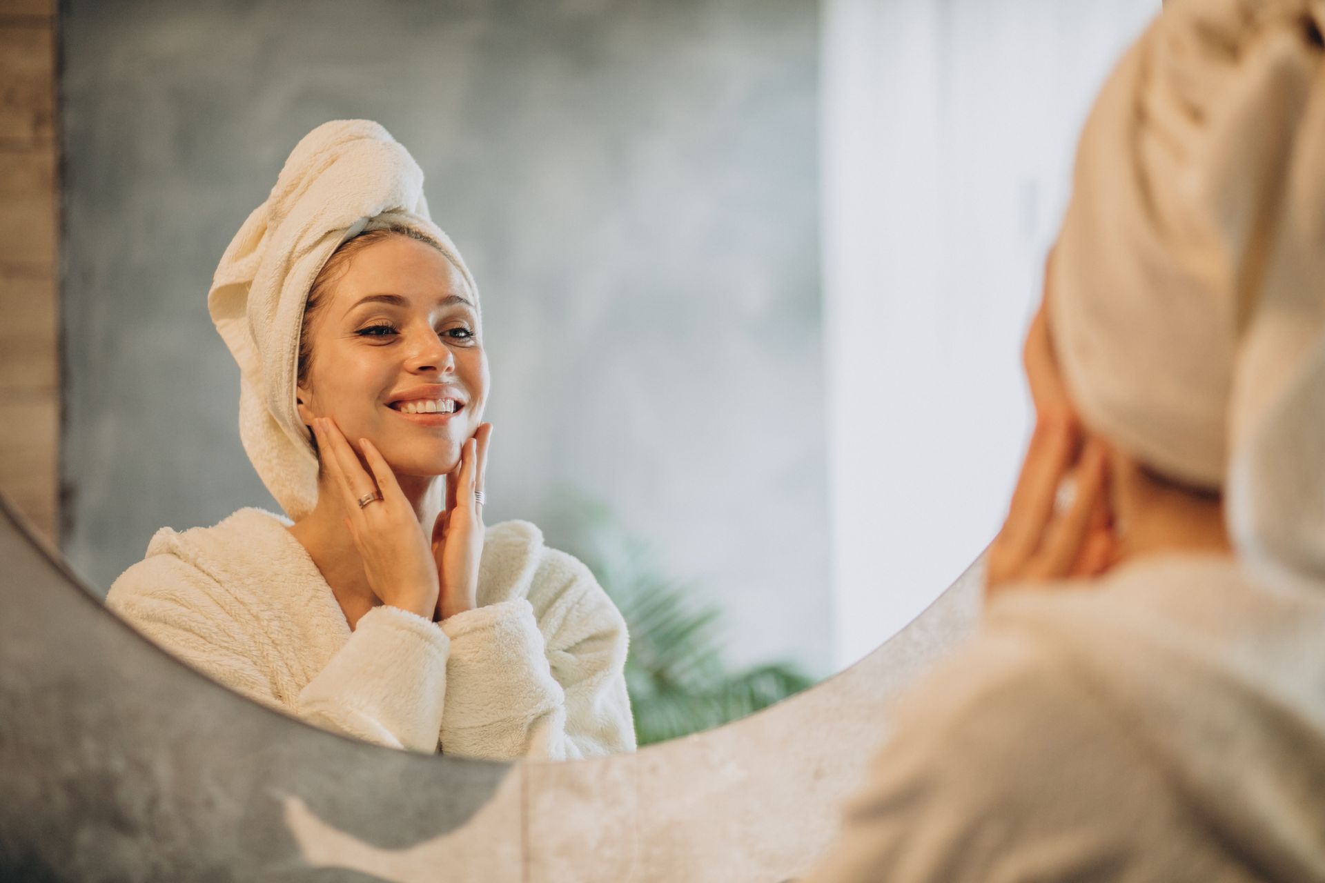Woman in robe and towel looks in the mirror, smiling, touching her face.