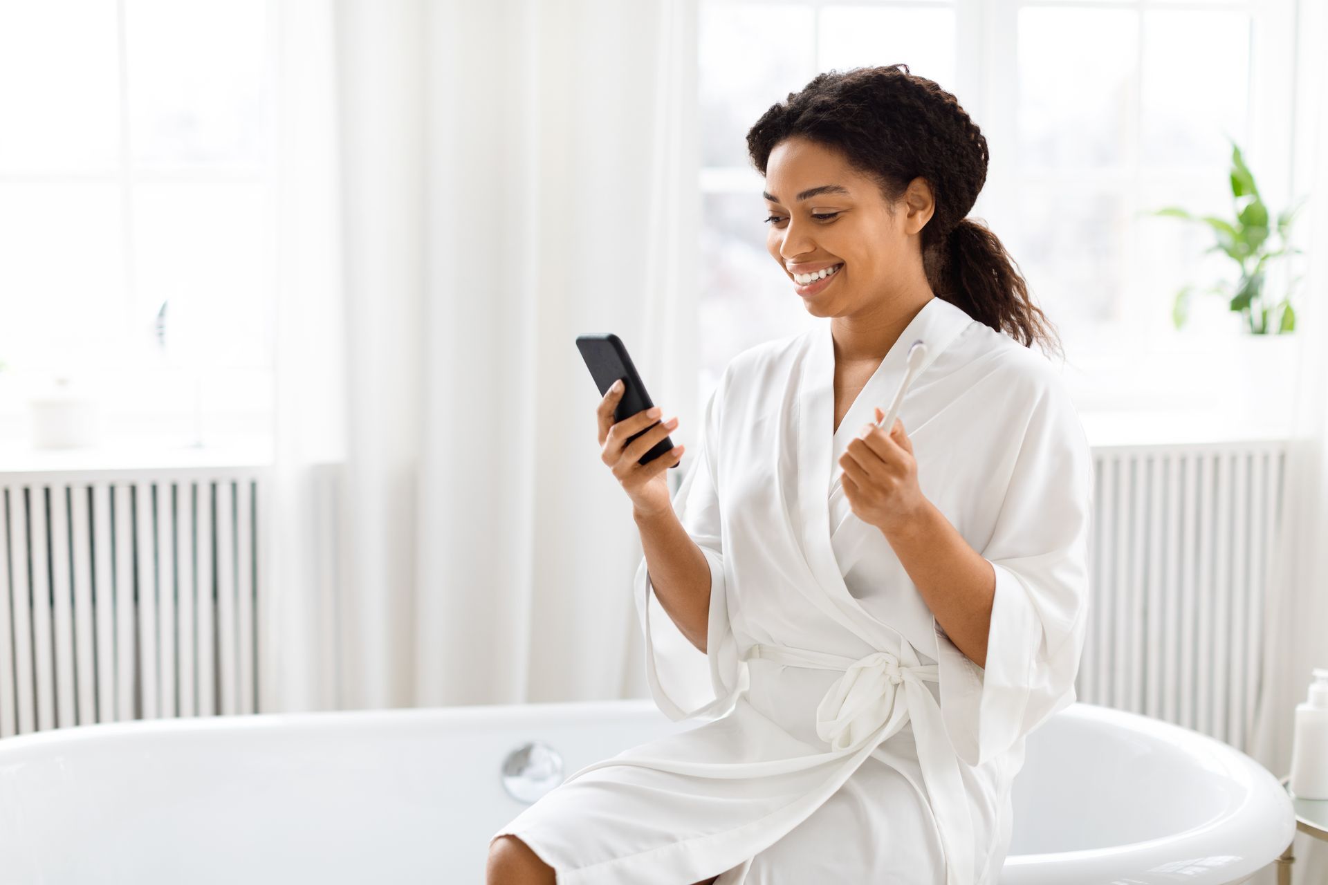 Woman in white robe smiles at her phone while sitting on a bathtub in a bright bathroom.