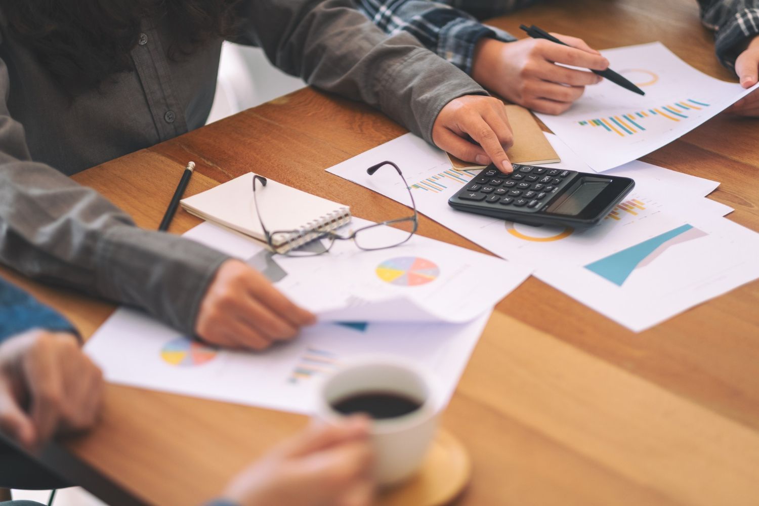 People reviewing financial documents at a wooden table, using a calculator and analyzing charts. Coffee cup present.