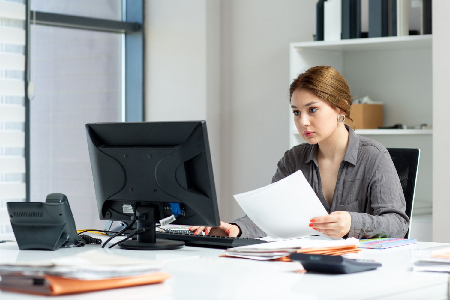 Woman in a gray shirt sits at a desk, looking at a computer screen and papers. Office setting.