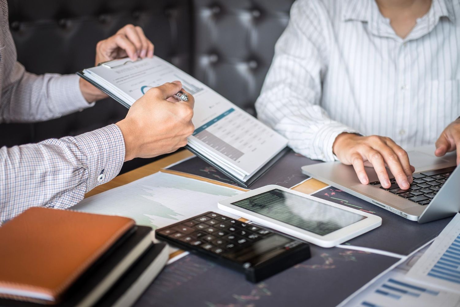 Two people reviewing financial documents, one using a laptop, with calculator, phone, and tablet on table.