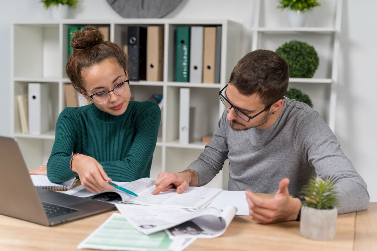 Two people in glasses reviewing documents at a desk with a laptop and shelves in the background.