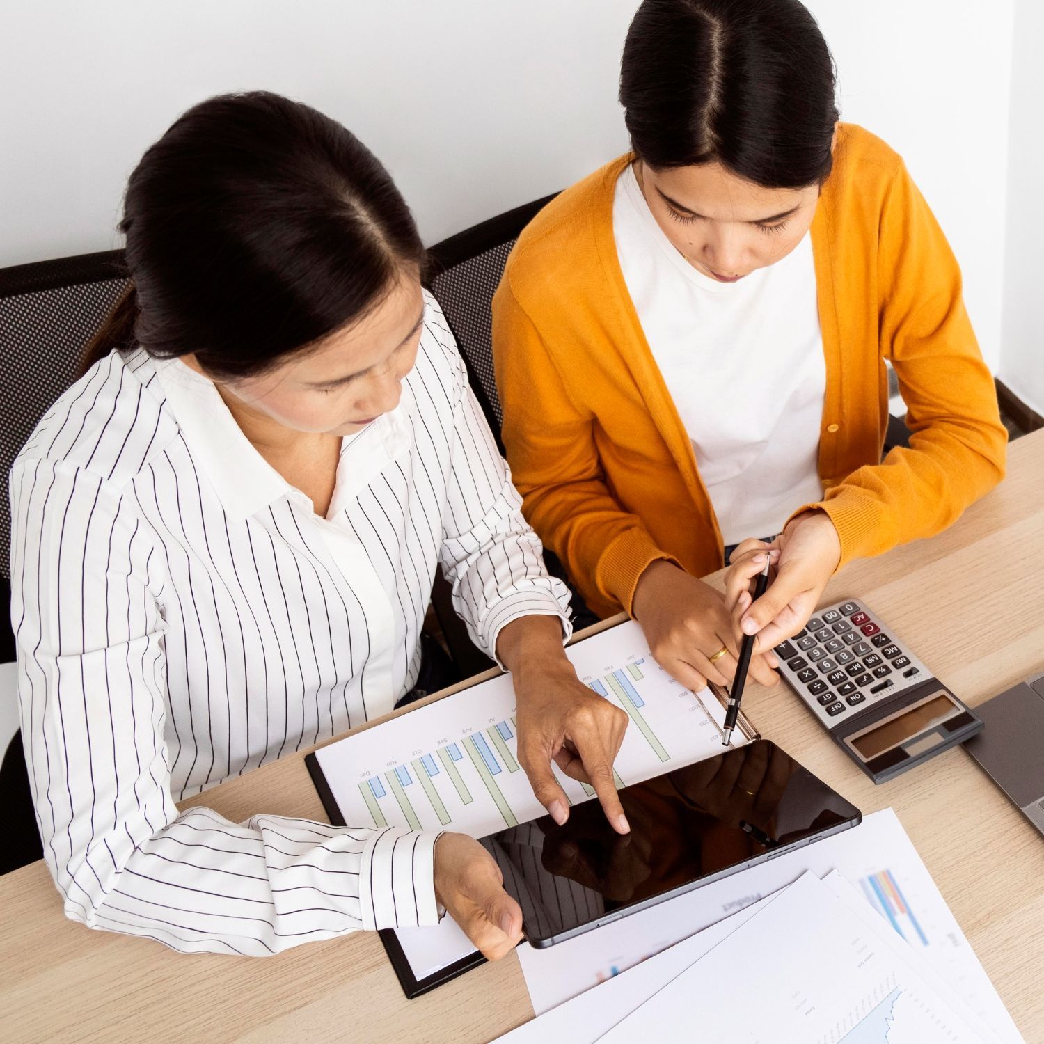 Two women working together, analyzing data on a tablet. One points, the other uses a pen near a calculator.