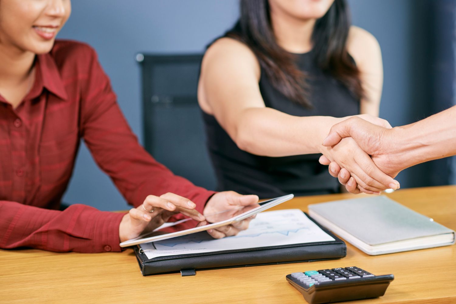 Two women at table with person shaking hands; one uses tablet, smiling.