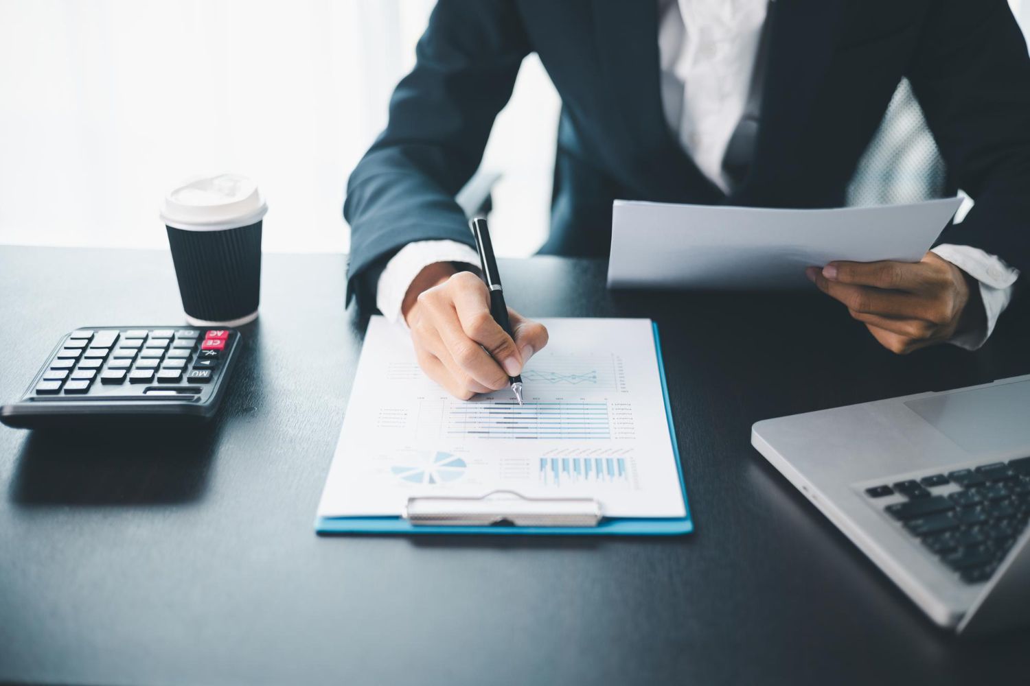 Person in suit reviewing documents at a desk with a calculator, laptop, and coffee.