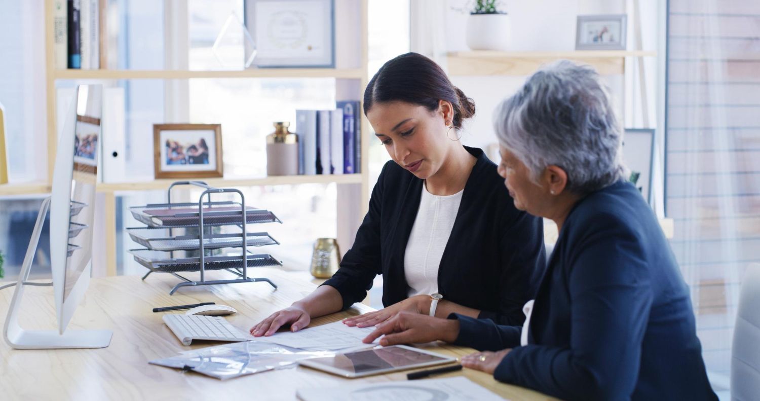 Two people at a desk review documents. One points at the paper; the other looks on.