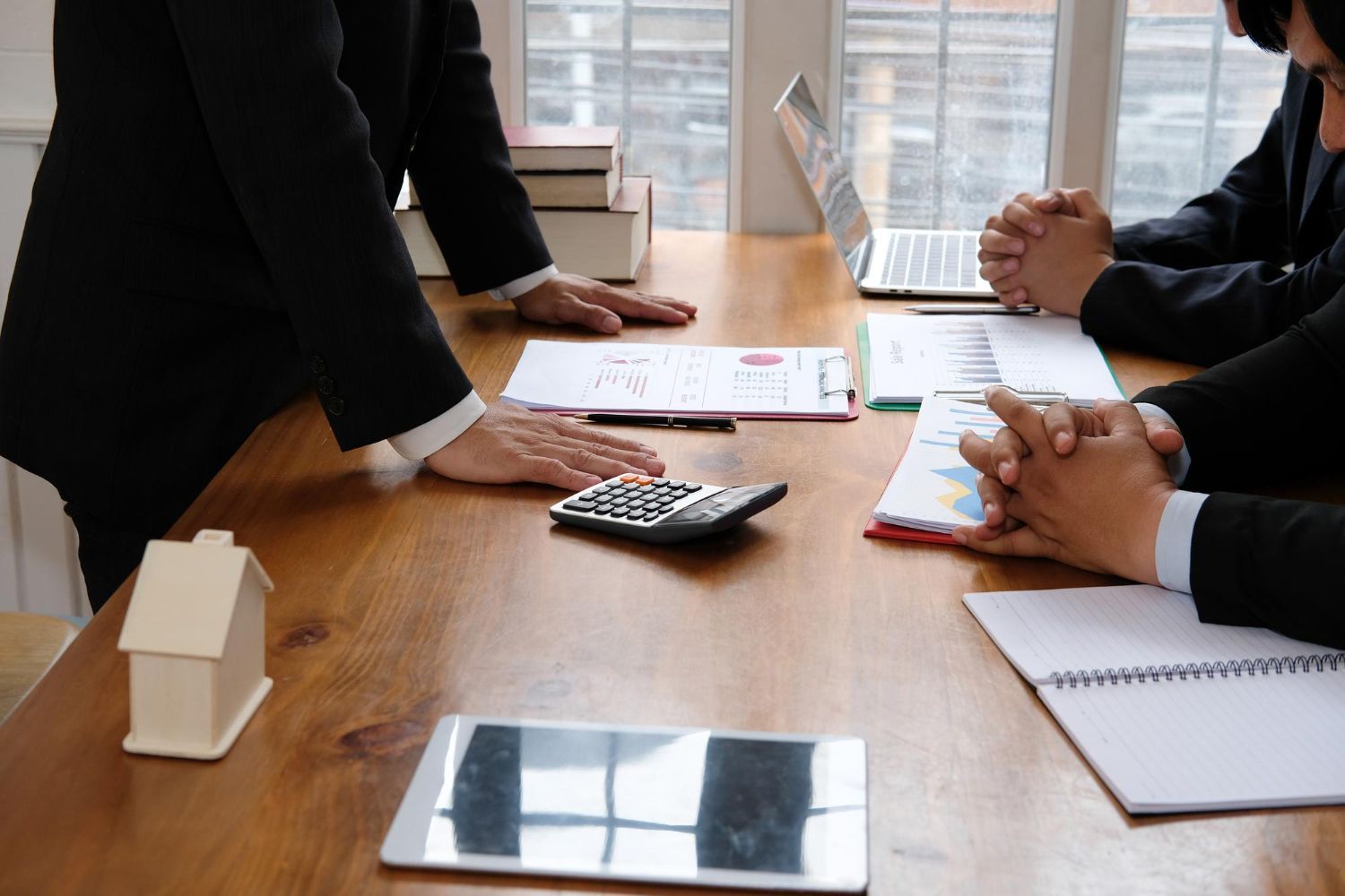 Business meeting: people in suits around a table with papers, laptop, calculator, and house model.