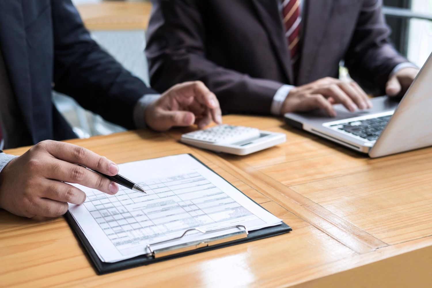 Two people in suits at a desk reviewing a document. One points with a pen, another uses a laptop.