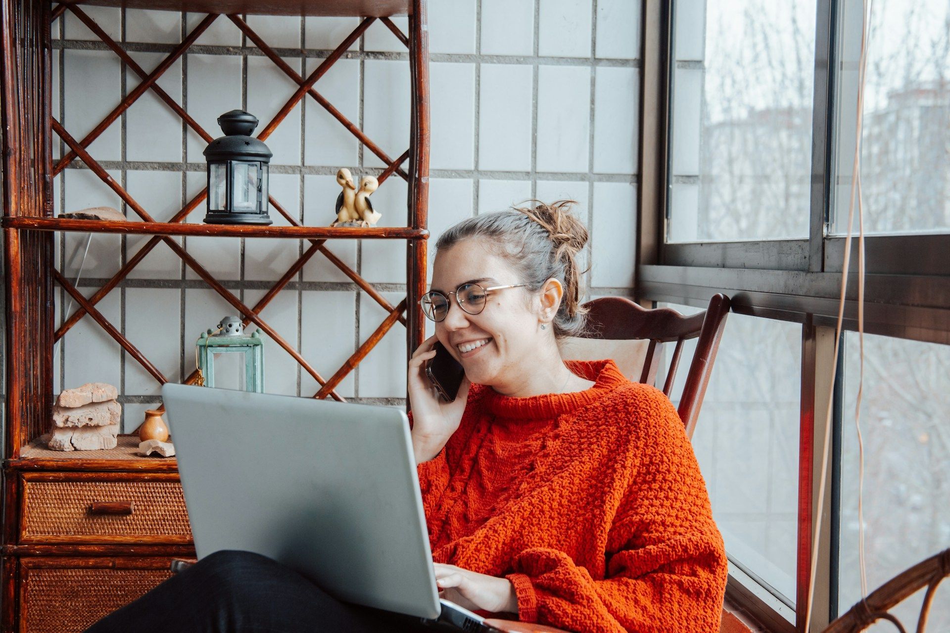 Woman with glasses smiles while on the phone, working on a laptop near a window.