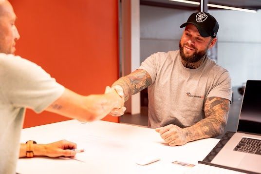 Twee mannen schudden elkaar de hand aan een bureau; een van hen heeft tatoeages en een Raiders-pet en glimlacht.