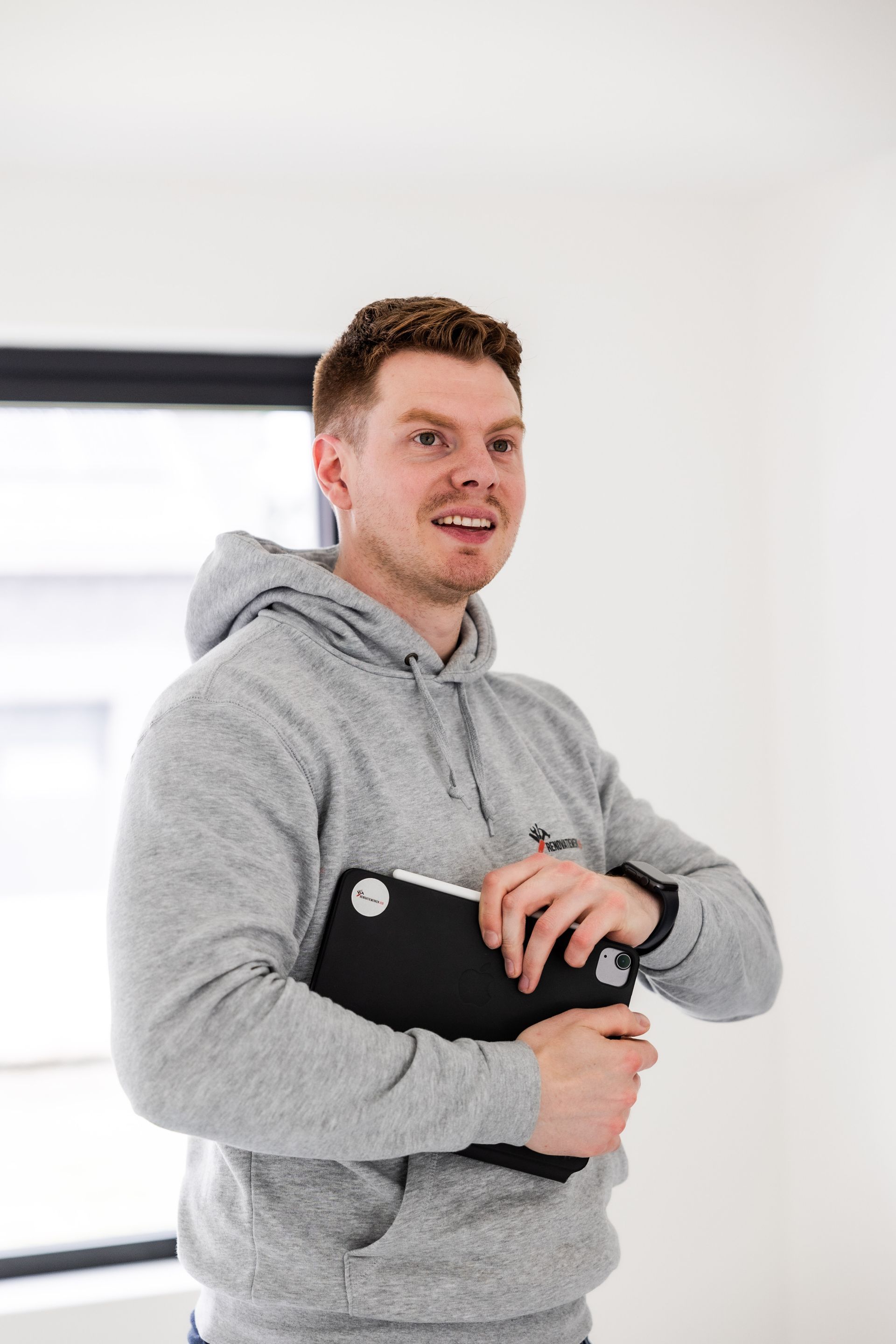 Man in grijze hoodie met zwarte weegschaal in zijn handen, lachend in een kamer met een raam.