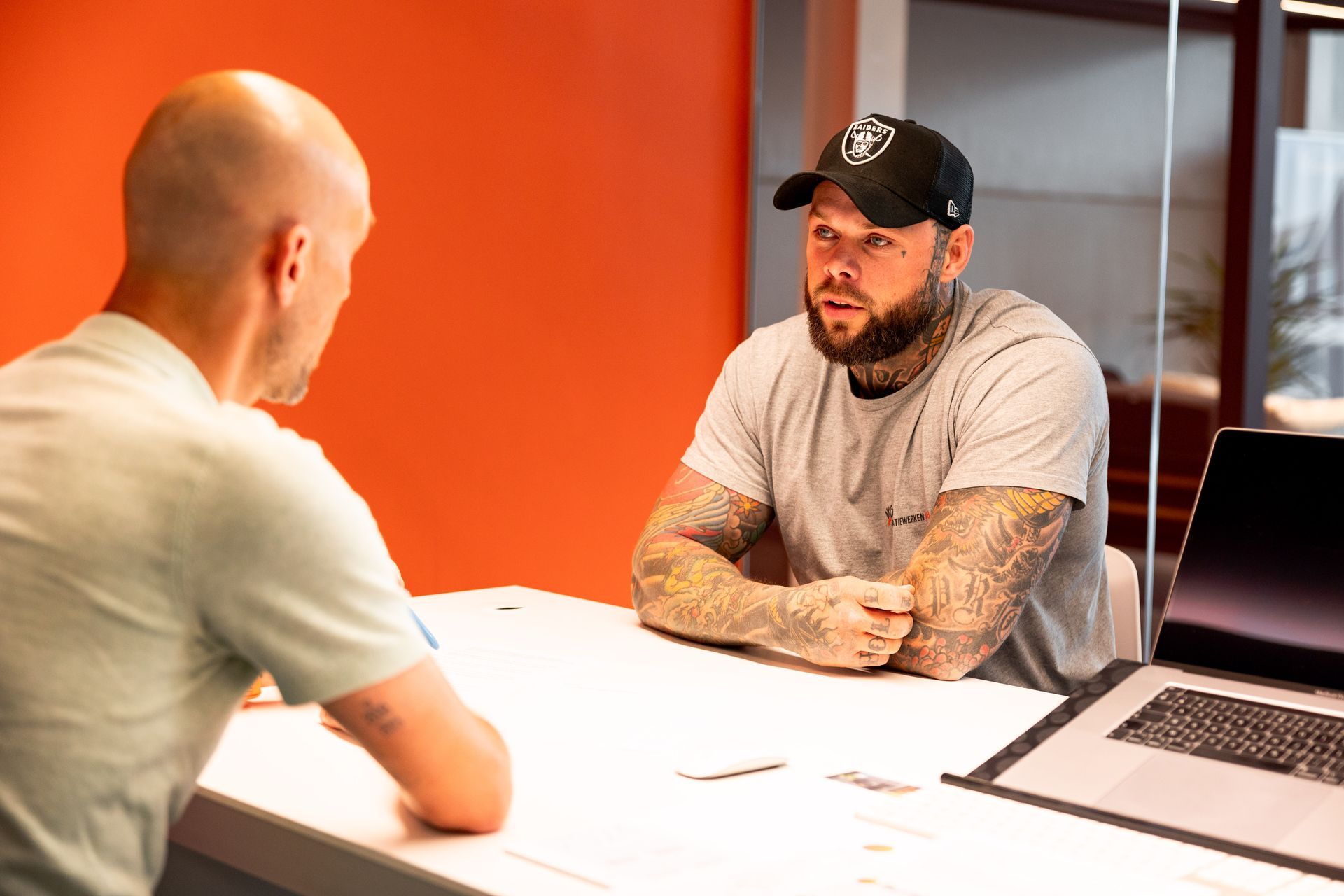 Twee mannen zitten aan een bureau, waarvan één een Raiders-pet draagt, met elkaar te praten in een kantoor met een oranje muur.