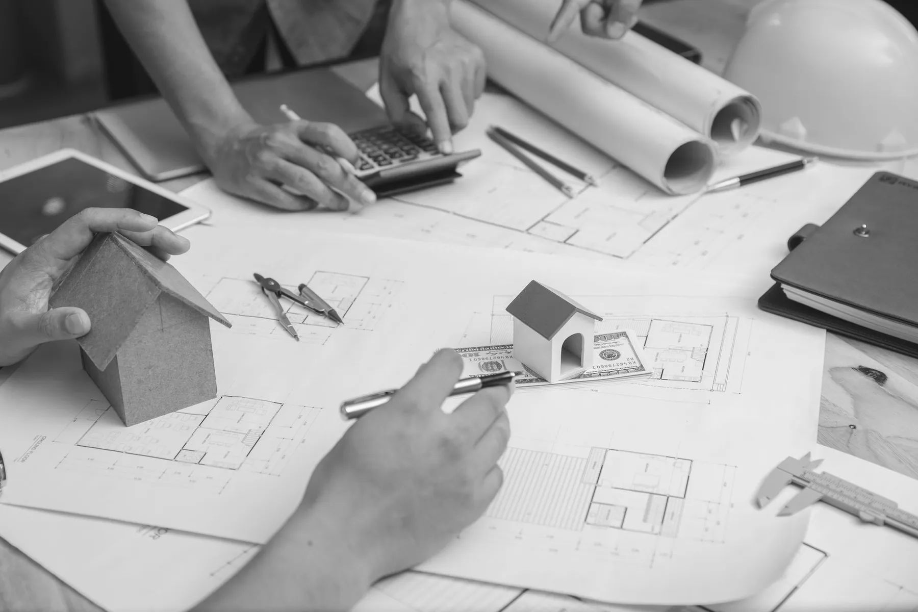 Handen die aan architectuurplannen werken met behulp van modellen, een rekenmachine en gereedschappen op een tafel.