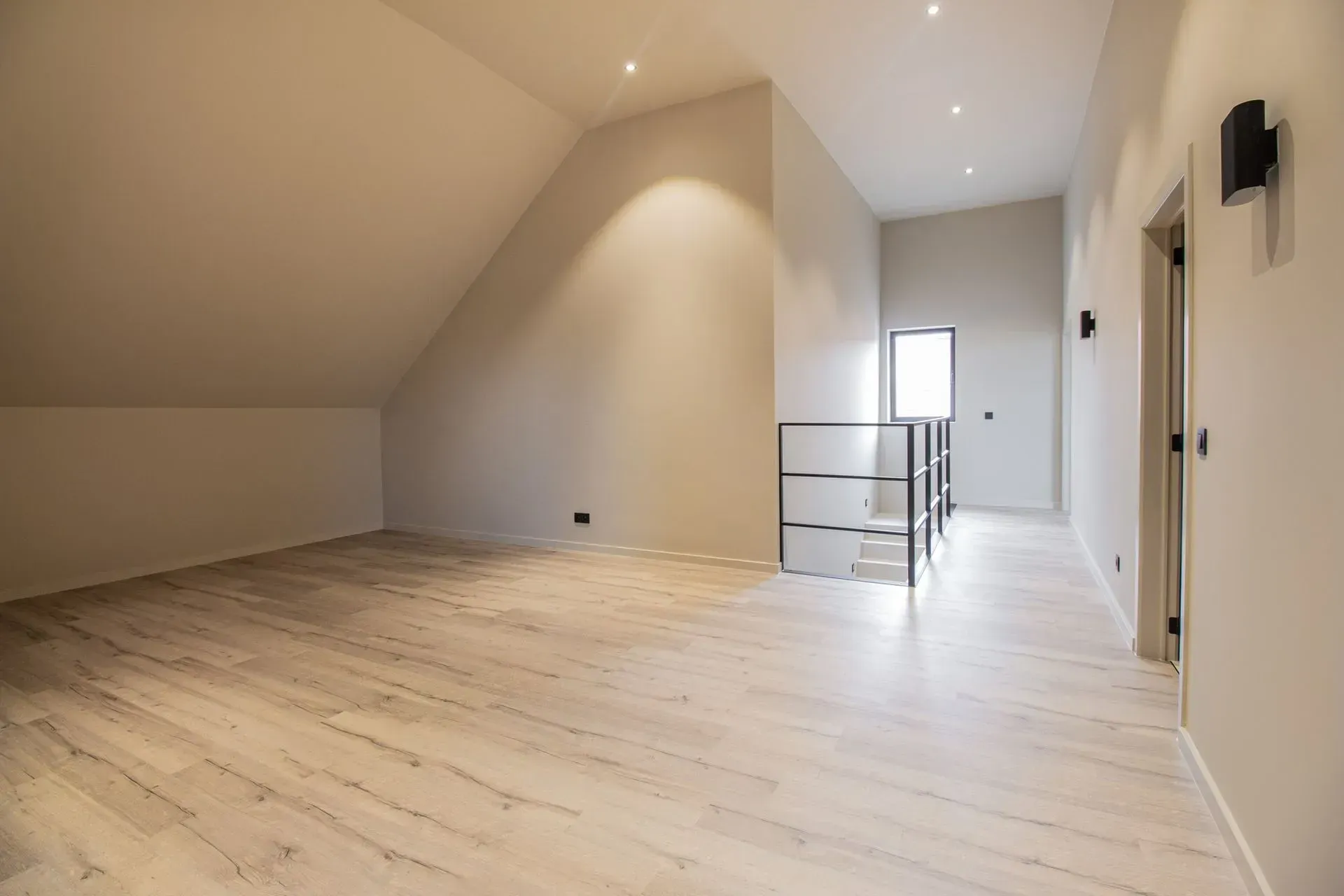 Lege zolderkamer met lichte houten vloer, schuin plafond en een zwarte metalen leuning.