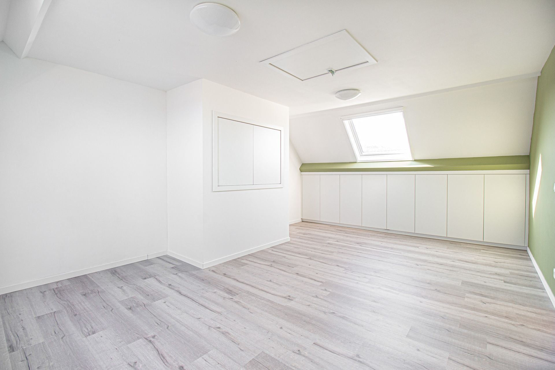 Lege kamer met witte muren, lichte houten vloeren en een klein raam; ingebouwde kasten langs een van de muren.