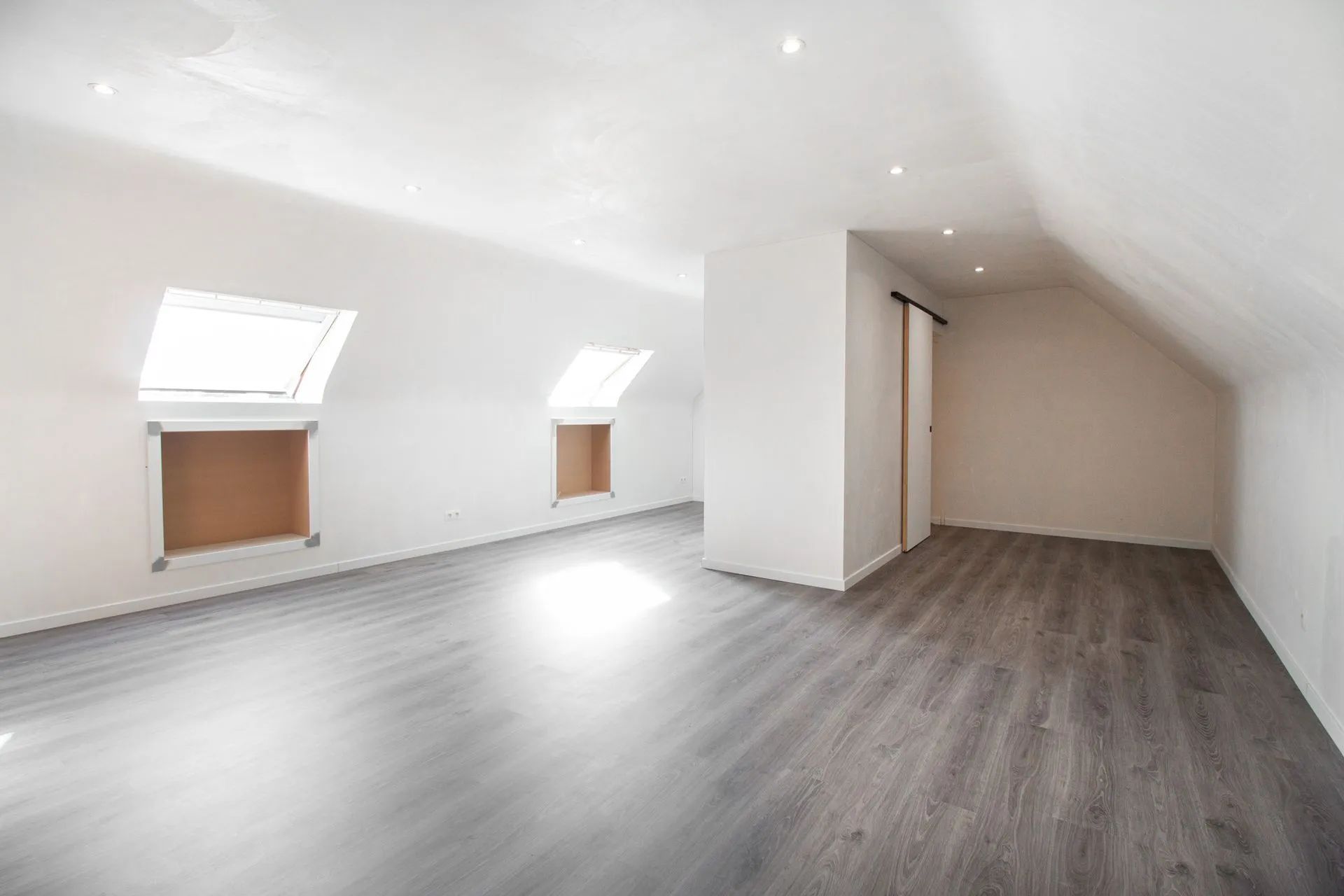 Lege, lichte zolderkamer met grijze houtlook vloer, schuine plafonds, drie ramen en een deur.